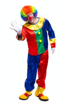Un homme vêtu d’un costume de clown rouge, bleu, vert et jaune nous fait signe. Il a une perruque multicolore, un maquillage blanc et un gros nez rouge